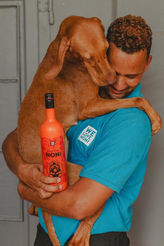 Smiling man in a blue We Love Noni shirt hugging a brown dog while holding an orange bottle of Dogs Love Noni.
